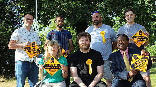 Three people sat on a bench in front of four people standing behind them. They are wearing Liberal Democrat rosettes and holding Liberal Democrat signs.